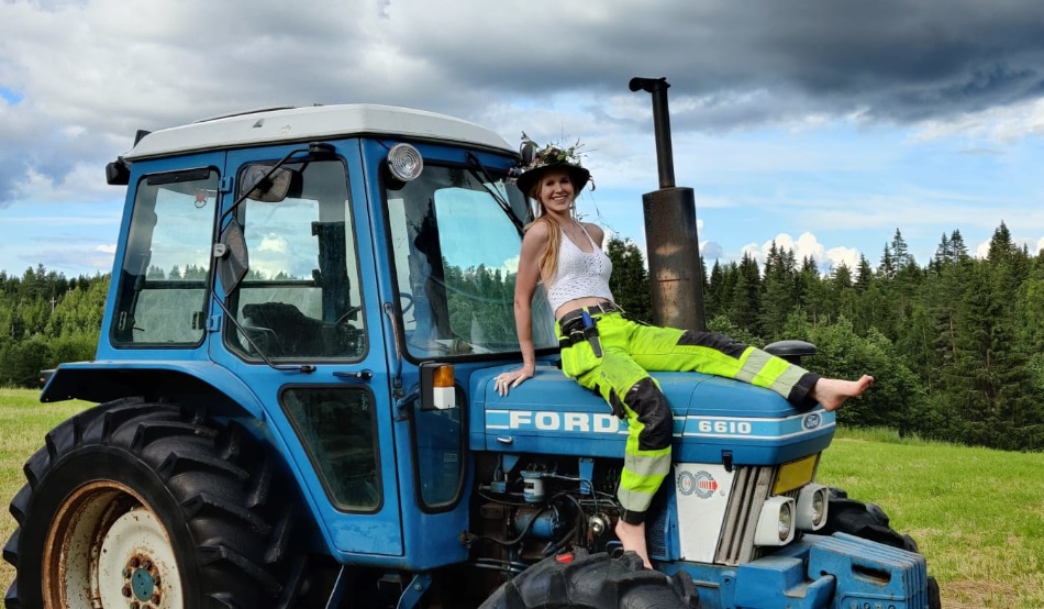 Traktorin konepellin päällä istuu nainen työhousuissa ja kukkaseppele päässä.