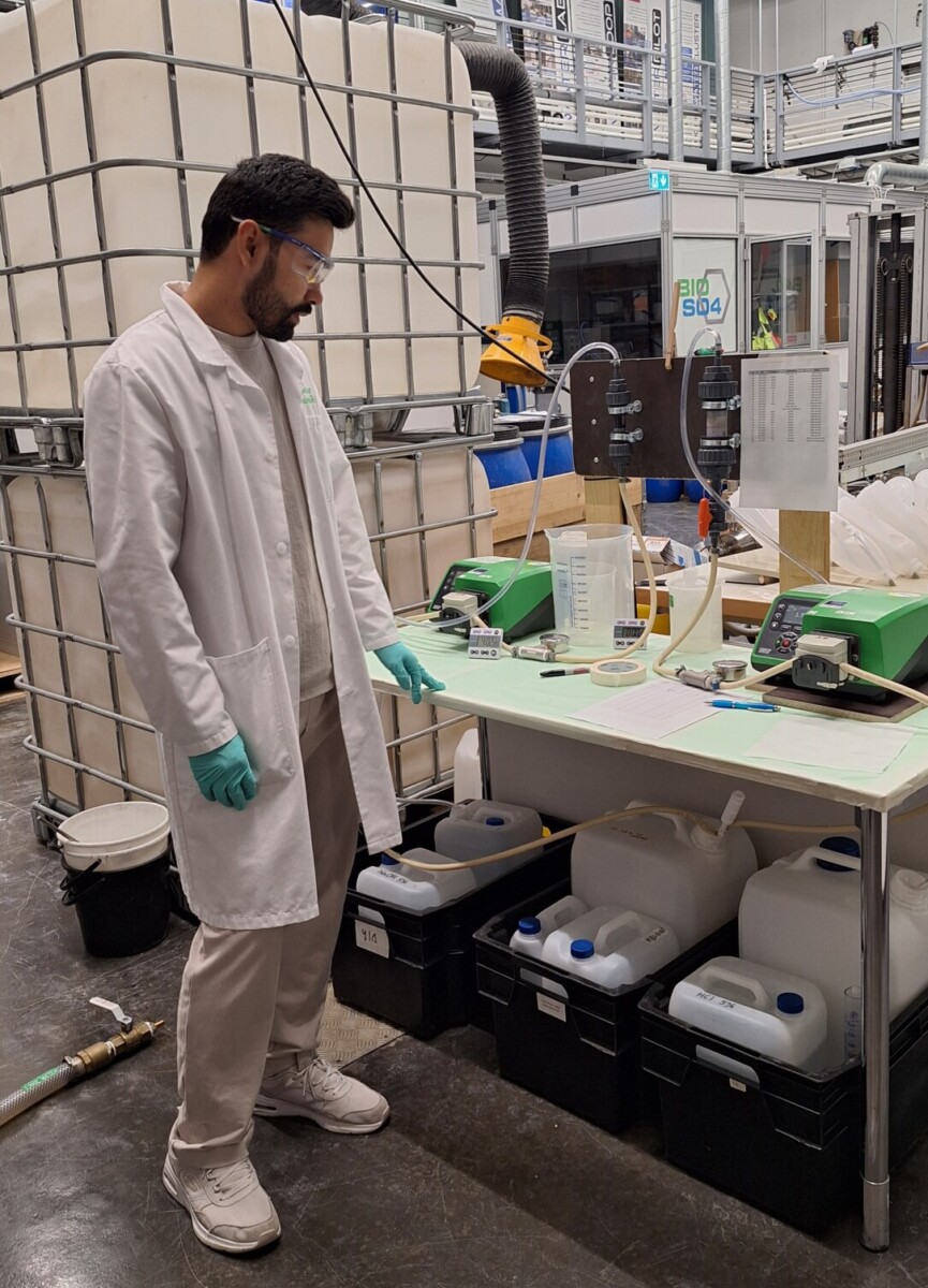 valkotakkinen mies seisoo tuotantotiloissa pöydän ääressä, jossa erilaisia mittauslaitteistoja.