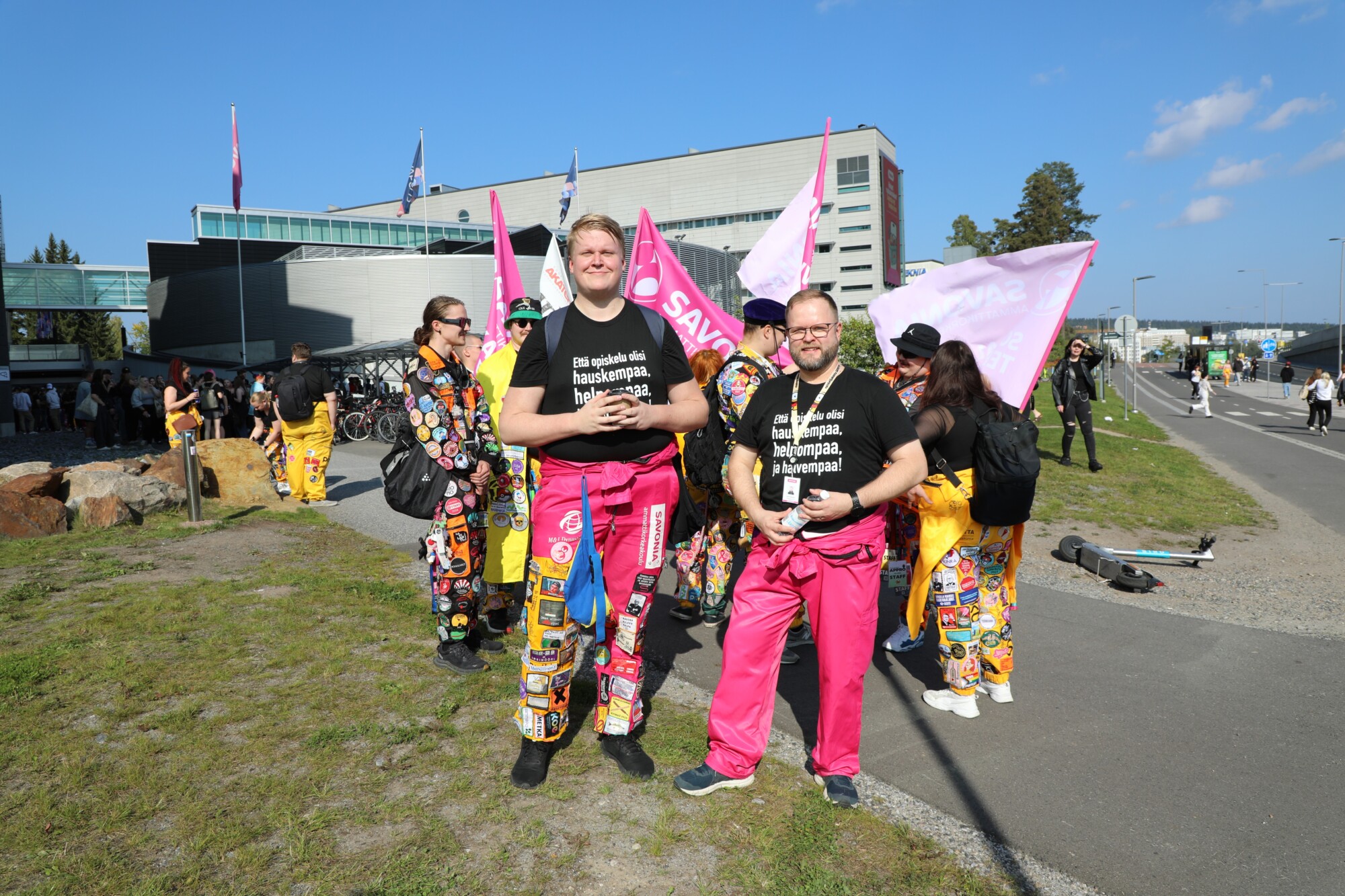 Oppilaskunnan puheenjohtaja ja toiminnanjohhtaja haalarit päällä ulkona kuvattuna, taustalla kampus ja haalaripukuisia opiskelijoita ja lippuja.